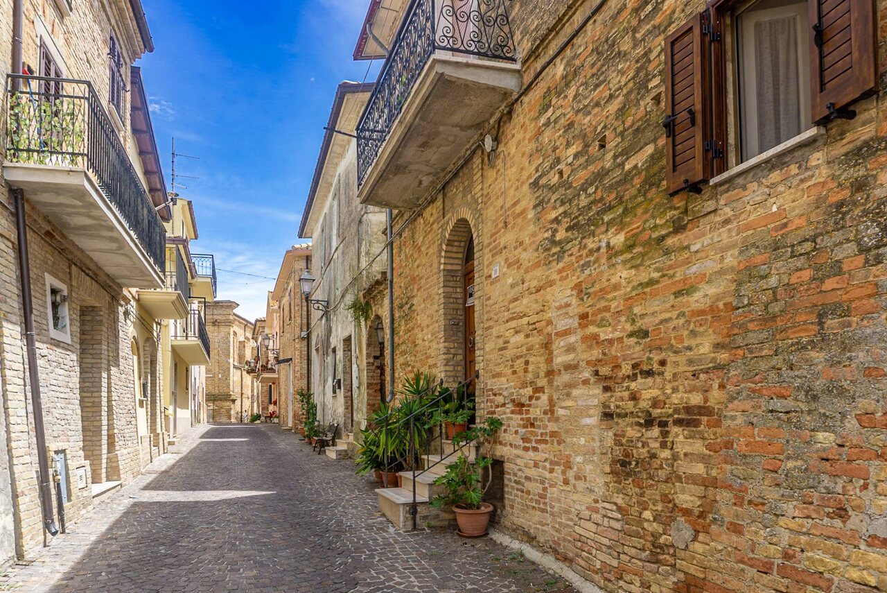 Vorher: Originalfoto einer Altstadtgasse in Italien bei Sonne, mit Backsteinhäusern, schmiedeeisernen Balkonen, trockenem Kopfsteinpflaster und hellblauem Himmel