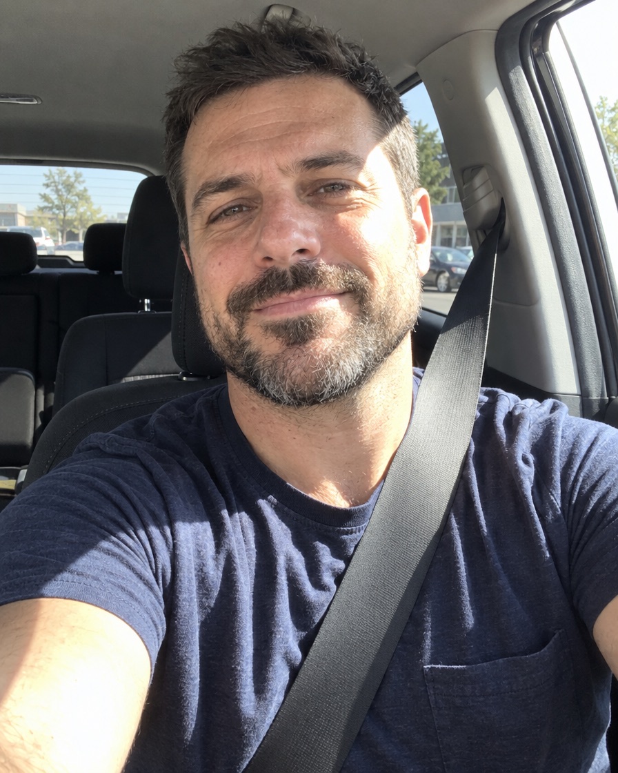 Derselbe Mann als Selfie im Auto mit Sicherheitsgurt und dunkelblauem T-Shirt, hartes Sonnenlicht wirft Schatten auf eine Gesichtshälfte