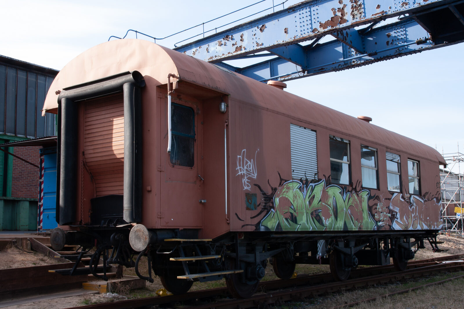 Brauner Reisezugwagen mit Graffiti auf einem Gleis im Hafen, darüber ein blauer Kranträger mit Rostflecken