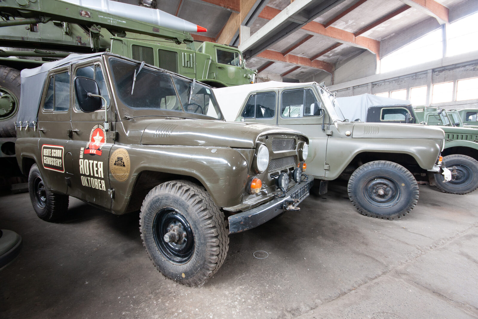 Olivgrüner offener Geländewagen mit Aufklebern in einer Halle voller Militärfahrzeuge, oben links eine Rakete