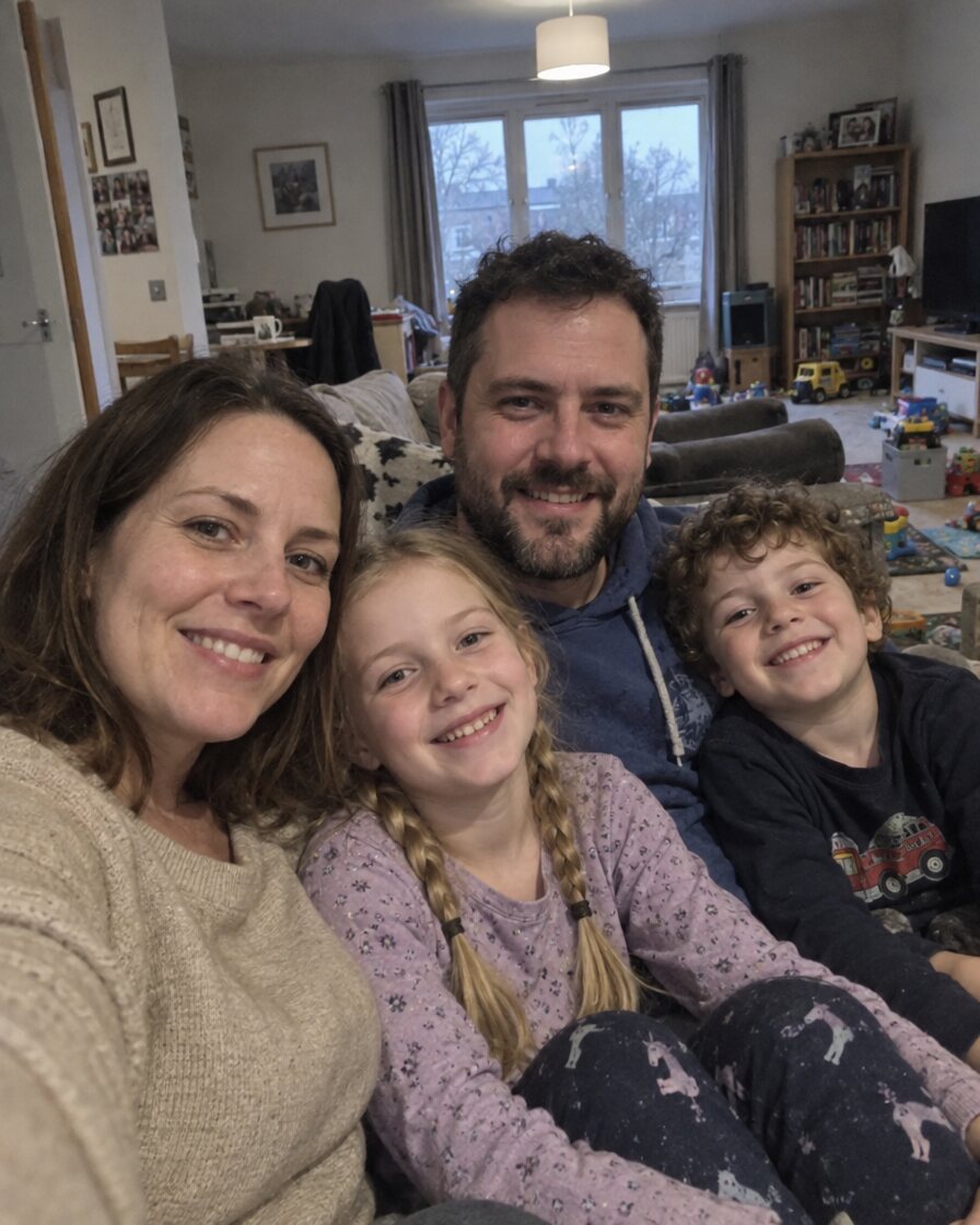 Handy-Selfie einer Familie mit zwei Kindern auf dem Sofa, graues Tageslicht, unaufgeräumtes Wohnzimmer
